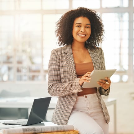 smiling professional woman holding a tablet and leaning on a desk in a bright office facing camera