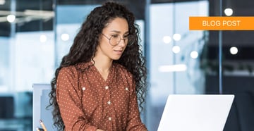 A woman with curly hair and glasses works on a laptop in a modern office, representing help desk managed services.