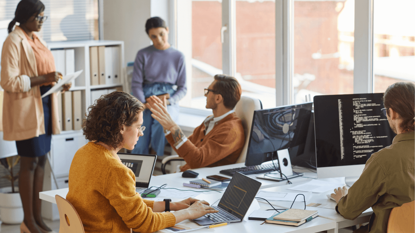 Several employees at work using computers while talking to each other.