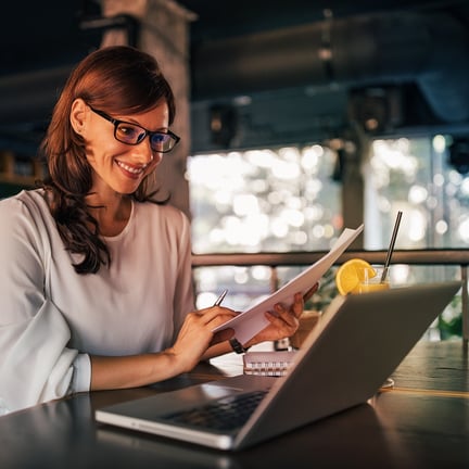 smiling woman in glasses seated at laptop in cafe holding paperwork