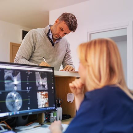 patient filling out paperwork in front of technician seated at dental office desk viewing dental xrays