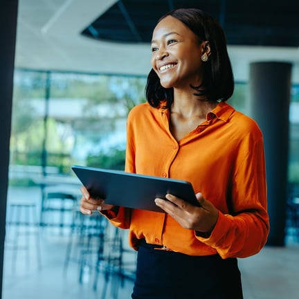 smiling business executive in natural light standing in modern office holding tablet looking out window
