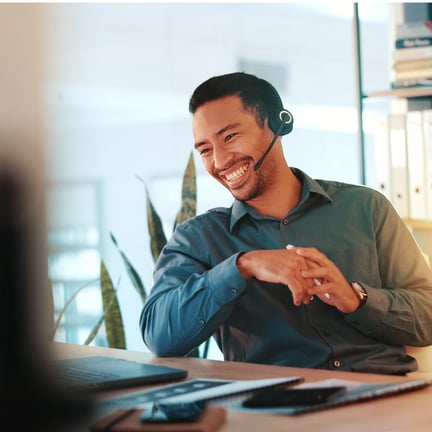 smiling man wearing headset speaking to client over the phone seated at laptop on desk