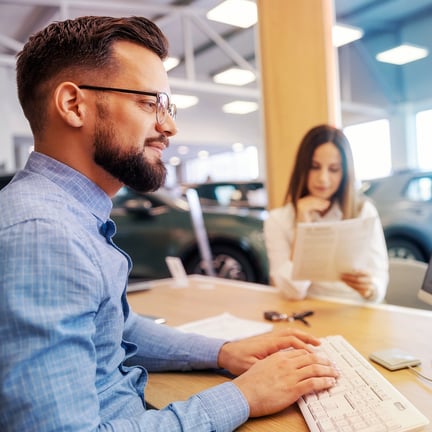 Smiling car salesperson working on computer while buyer reads contract at automobile dealership