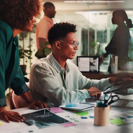 two business professionals in bustling office discussing ideas over laptop