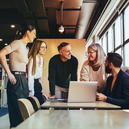 five smiling business professionals collaborating around a laptop in a bright office.