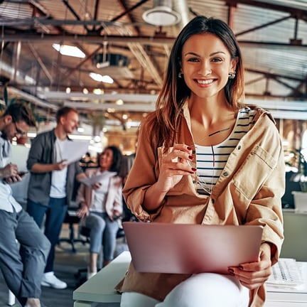 smiling young woman holding a laptop and glasses, seated in a busy, modern open office with her coworkers in the background