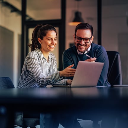 two smiling colleagues collaborating on a laptop at a dark conference table